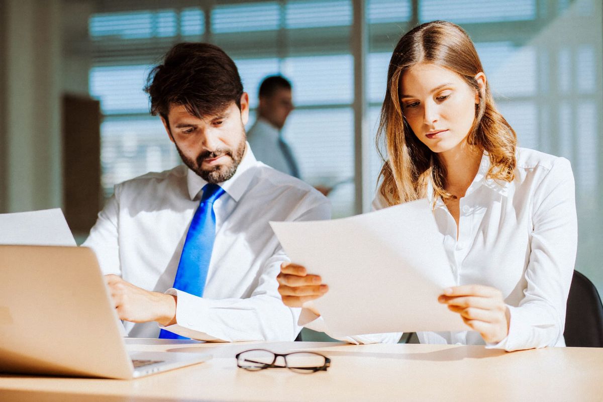 Dos profesionales revisando documentos impresos junto a una laptop, en referencia a la aplicación consistente de reglas de auditoría